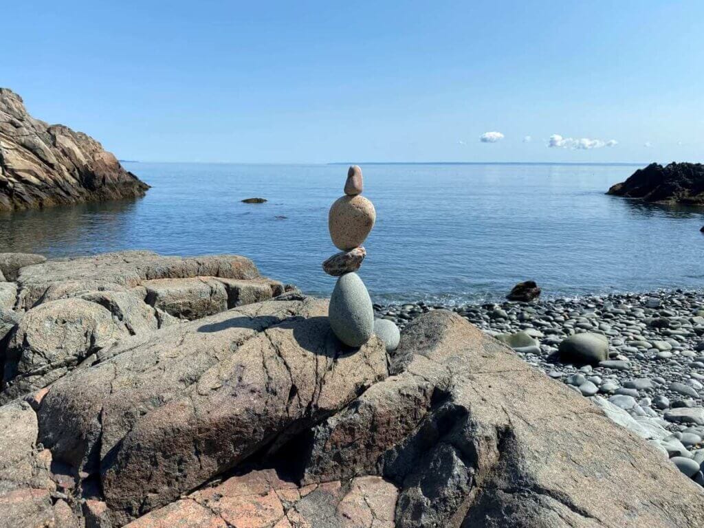Stack of four smaller rocks balancing on larger rock on rocky beach with ocean in background